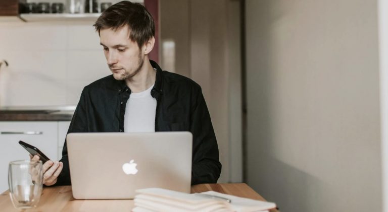 Man working on laptop while holding smartphone.
